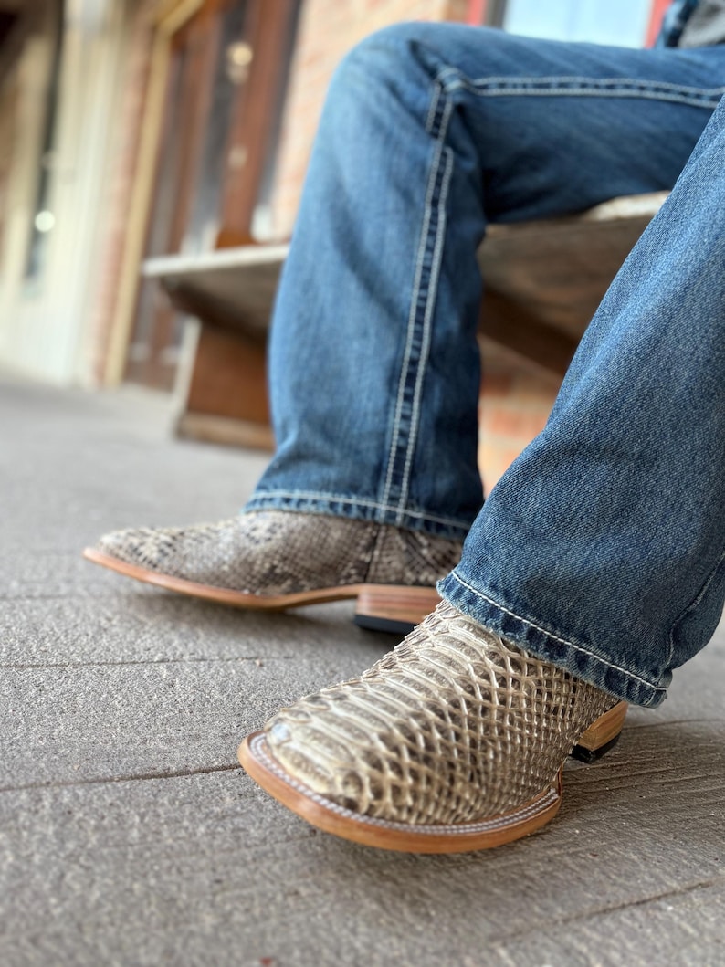 Peut inclure: Gros plan sur une paire de bottes de cowboy &agrave; motif peau de serpent. Les bottes ont un bout carr&eacute; et une semelle marron clair. La personne porte un jean bleu.