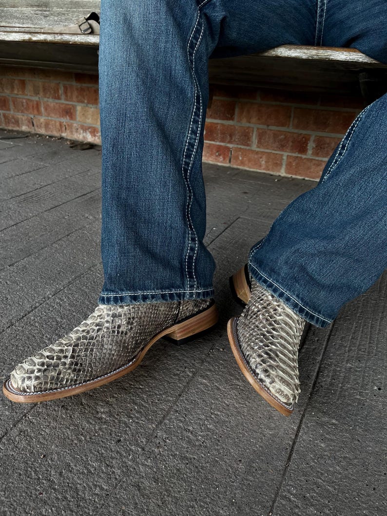 Peut inclure: Bottes de cowboy en peau de serpent avec un bout carr&eacute; et des semelles marron clair. Les bottes sont port&eacute;es avec un jean bleu. Les bottes ont une palette de couleurs neutres de gris, blanc et marron.