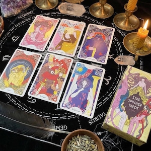 May include: A collection of tarot cards with colorful illustrations, arranged on a black surface with astrological symbols. A box labeled "Star Spinner Tarot" is visible, along with candles, crystals, and a feather. The cards feature various figures and designs.