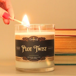 May include: A clear glass jar candle with white wax and two wicks being lit by a match. The label is black with gold text reading Plot Twist, Aged Papyrus, Birch & Cedar Wood, Leather, and Hand Poured in Virginia, with a net weight of 9oz.