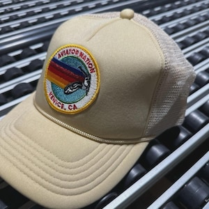 May include: Tan trucker hat with a mesh back and a round patch. The patch features the text "AVIATOR NATION" and "VENICE, CA" around a colorful graphic. The hat has a curved brim and a rope detail.
