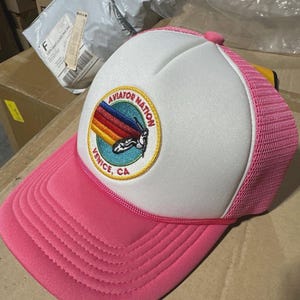 May include: Pink and white trucker hat with a curved brim. The hat features a patch with the text "AVIATOR NATION VENICE, CA" and a rainbow design. The hat has a pink mesh back and a pink button on top.