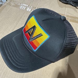 May include: Black trucker hat with a structured front and mesh back. The hat features a rectangular patch with a colorful design of a gradient of yellow, orange, and red, with a black letter "A" in the center. The patch is outlined in blue.