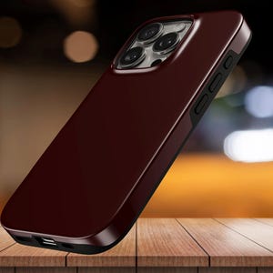 May include: A burgundy phone case with a glossy finish. The case has a black border and features a camera cutout with multiple lenses. The phone case is displayed on a wooden surface.