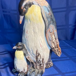 May include: A ceramic penguin sculpture featuring a large adult penguin and a smaller chick. The adult penguin has a black head, yellow chest, and a white belly. The sculpture is painted with muted colors and has a textured appearance.