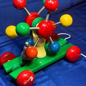 May include: A colorful wooden pull toy featuring a green base with red wheels and a central structure of red, yellow, green, and blue spheres connected by springs. The toy has a white pull string.