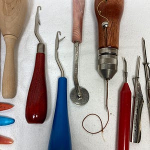 May include: A collection of vintage craft tools, including wooden handles, metal hooks, and a pink-handled rotary cutter. The tools feature red, blue, and brown handles, and are arranged on a white surface. Ideal for crafting and DIY projects.