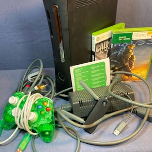 May include: A black Xbox 360 game console with a translucent green wired controller, a power brick, a black wireless network adapter, and two game cases, Call of Duty Black Ops and Halo 3, along with a small white instruction manual and cables.