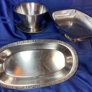 May include: A set of stainless steel serving dishes. The set includes a gravy boat with a saucer, a rectangular dish, and an oval platter with a decorative border. The items are displayed on a blue fabric background.