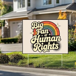 May include: A yard sign with the text "Big Fan of Human Rights" in a retro font, featuring a rainbow and starburst design. The sign is displayed on a green lawn in front of a house.