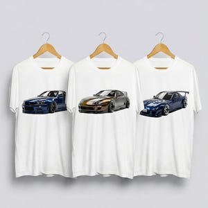 Puede incluir: Tres camisetas blancas colgadas en perchas de madera, cada una con un diseño de coche diferente. Los coches son azul oscuro, bronce y plata, y un coche deportivo azul. Las camisetas son blancas.