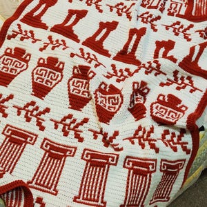 May include: A white and red crochet blanket featuring a repeating pattern of Greek columns, vases, and decorative elements. The blanket has a red border and is made with a textured stitch, creating a cozy and visually appealing design.