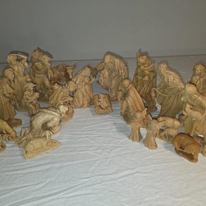 以下が含まれることがあります： 木彫りの降誕のフィギュアのコレクション。マリア、ヨセフ、幼子イエス、羊飼い、動物が含まれます。フィギュアはさまざまなポーズで、顔の特徴や衣服が詳細に表現されています。セットは白い表面に表示されています。