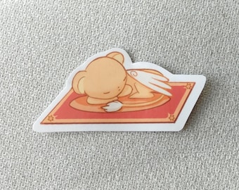 May include: A sticker featuring a cartoon character with a bear-like appearance, wings, and a sleeping pose. The character is light brown and rests on a red and orange rug with star-shaped corner designs. The sticker has a white border.