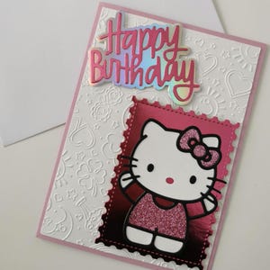 May include: A handmade birthday card with a Hello Kitty design. The card has a white embossed background with hearts and floral patterns. The words "Happy Birthday" are in pink and iridescent lettering. The Hello Kitty image is in a pink frame.