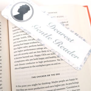 May include: A rectangular bookmark with the text "Dearest Gentle Reader" in a script font. The bookmark features a silhouette of a woman in a circle at the top. The bookmark is placed in an open book.
