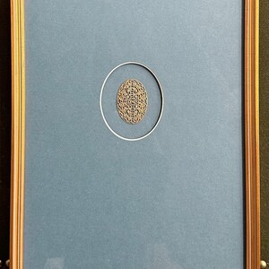 以下が含まれることがあります： くすんだ青色の背景に、金色の複雑な模様の楕円形のデザインが施された額装されたアートワーク。 アートワークは金色の木製フレームに額装されており、クラシックでエレガントな美しさを生み出しています。 楕円形は細い銀色のリングで囲まれています。