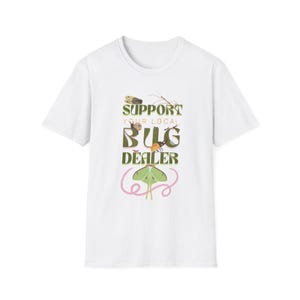Può includere: Una t-shirt bianca a maniche corte con una stampa grafica che riporta il testo SUPPORT YOUR LOCAL BUG DEALER in lettere verde scuro. Il design include illustrazioni di una cicala, un insetto stecco, una lumaca, uno scarabeo e una grande falena luna verde.