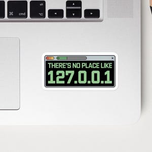 May include: A sticker with the text "THERE'S NO PLACE LIKE 127.0.0.1" in a retro computer window design. The sticker is on a laptop, with a black smartphone, a pencil, a pen, and a red notepad nearby.
