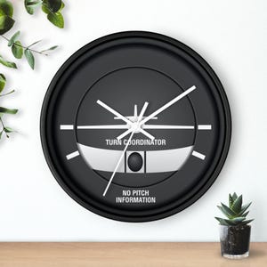 May include: A round black wall clock featuring a white graphic of an aircraft turn coordinator instrument. The face displays the text TURN COORDINATOR and NO PITCH INFORMATION in white, with white clock hands indicating the current time.