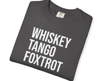 May include: A charcoal gray t-shirt featuring the white text WHISKEY TANGO FOXTROT printed in a bold, sans-serif font across the chest. The garment has a crew neckline and a visible interior brand label at the collar area, folded neatly.