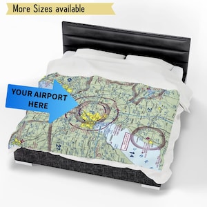 Puede incluir: Una manta con un diseño detallado de mapa de aviación, con una flecha azul que apunta a "YOUR AIRPORT HERE". La manta está sobre una cama con cabecero negro y ropa de cama blanca. El texto en la parte superior dice "More Sizes available."