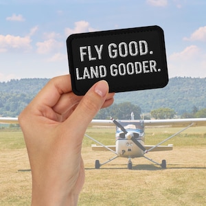 May include: Black rectangular patch with white embroidered text reading "FLY GOOD. LAND GOODER." held in front of a small airplane on a grassy field. The sky is blue with scattered clouds.