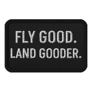May include: Black rectangular patch with white embroidered text that reads "FLY GOOD. LAND GOODER." The patch has a stitched border and a textured appearance, suitable for application on clothing or gear.