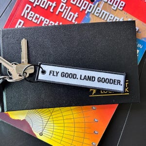 May include: A keychain with keys and a rectangular tag. The tag is light blue with the text "FLY GOOD. LAND GOODER." in black. The keychain is on top of a black notebook and other printed materials.