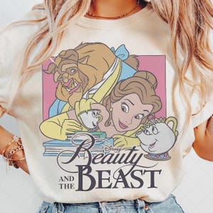 May include: Cream-colored t-shirt featuring a graphic of Belle and the Beast from Disney's Beauty and the Beast. The design includes the characters, teacups, and the movie title in a stylized font.