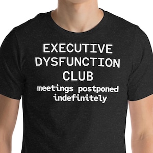 May include: Dark grey t-shirt featuring the white text "EXECUTIVE DYSFUNCTION CLUB" and below, "meetings postponed indefinitely". The t-shirt has a crew neck and is made of a soft material.