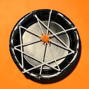 May include: A round black paper plate featuring a white yarn web pattern stretched across the rim. A small, bright orange plastic spider is positioned in the center of the web, creating a high-contrast decorative piece for seasonal displays.