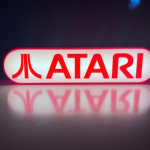 May include: A retro-style Atari sign, rectangular with rounded edges, glows brightly. The word "ATARI" is in bold red letters against a white background. The Atari logo is on the left side. The sign is reflected on a glossy surface.