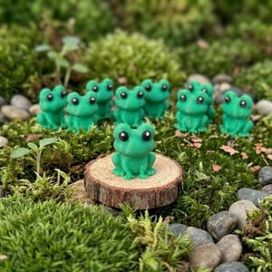 Könnte beinhalten: Eine Ansammlung kleiner, leuchtend grüner Froschfiguren mit schwarzen Augen ist auf einem Bett aus Moos, kleinen Steinen und Holzscheiben angeordnet. Ein Frosch sitzt auf einer Holzscheibe, während andere verstreut sind. Die Szene erinnert an einen Miniaturgarten.