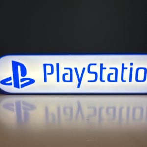 May include: A rectangular, illuminated sign displaying the word "PlayStation" in blue, with the PlayStation logo on the left. The sign has a white background and a blue border. The sign is reflecting on a white surface.