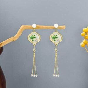 May include: Gold-toned dangle earrings featuring a white, cloud-shaped design with green bamboo accents and a small clear gem. The earrings have a pearl at the top and three dangling pearl beads at the bottom. The earrings are displayed on a wooden branch.
