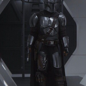 May include: A full-body shot of a character in a silver and black Mandalorian armor suit. The helmet is a polished silver, and the armor plates are detailed. The character is holding a blaster pistol. The background is a futuristic interior.