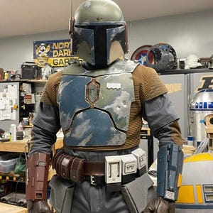 May include: A full-body costume featuring a weathered green helmet with a rangefinder, a blue and silver chest plate, a brown textured undershirt, and gray trousers. The outfit includes a brown utility belt with pouches and two distinct gauntlets.