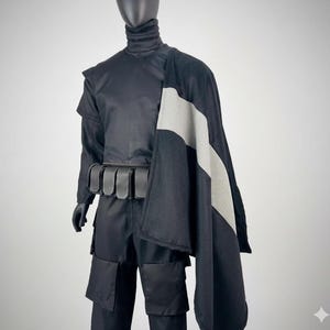 Peut inclure: Un costume noir présenté sur un mannequin. Il comprend un haut à col montant, un pantalon et une ceinture à poches. Une cape noire et grise est drapée sur l'épaule. Le costume est de style futuriste ou fantastique.