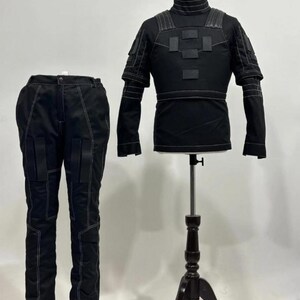 May include: A black tactical-style outfit consisting of a long-sleeved shirt with a high collar, shoulder padding, and rectangular chest patches, paired with matching trousers featuring reinforced knee panels and prominent white contrast stitching.