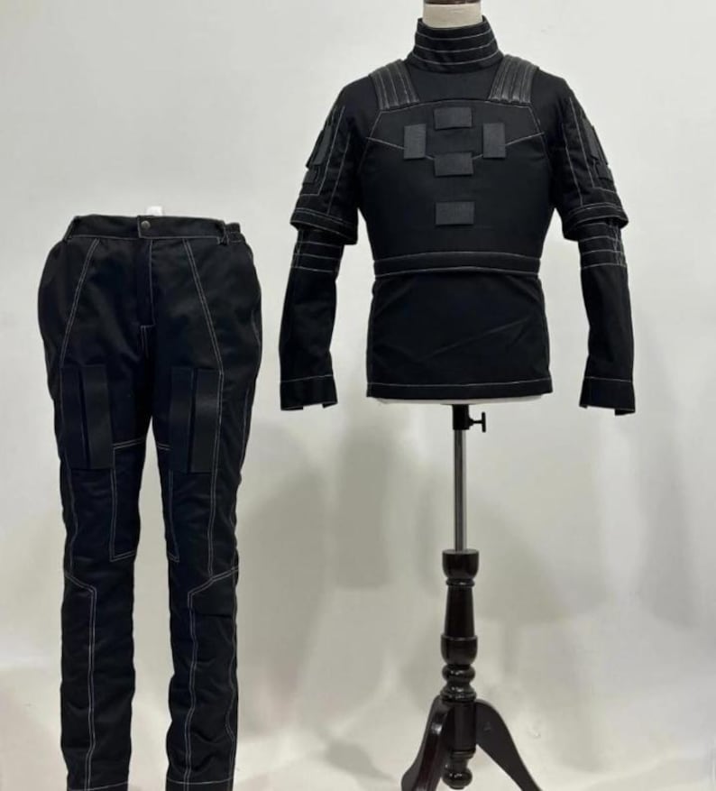 May include: A black tactical-style outfit consisting of a long-sleeved shirt with a high collar, shoulder padding, and rectangular chest patches, paired with matching trousers featuring reinforced knee panels and prominent white contrast stitching.