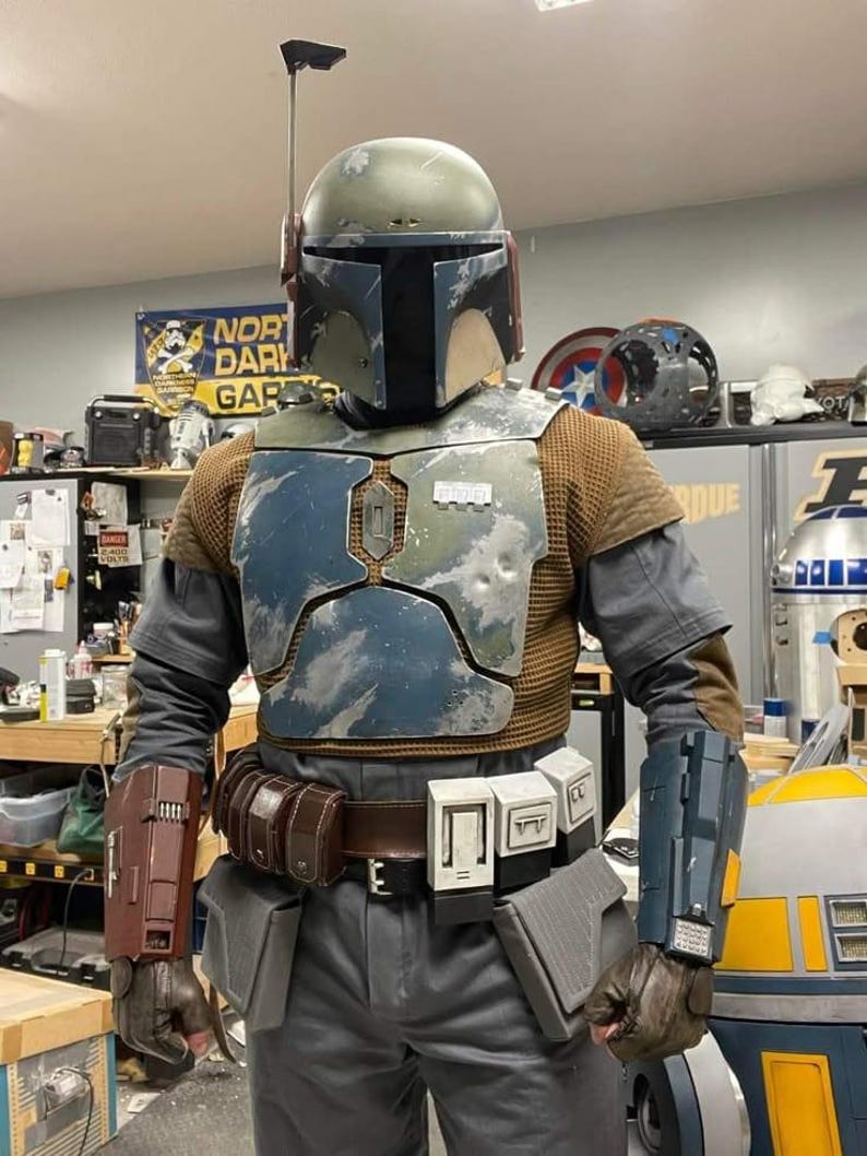 May include: A full-body costume featuring a weathered green helmet with a rangefinder, a blue and silver chest plate, a brown textured undershirt, and gray trousers. The outfit includes a brown utility belt with pouches and two distinct gauntlets.