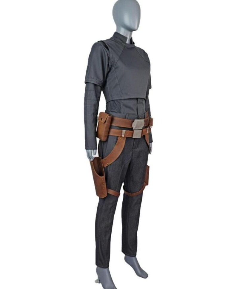 May include: A dark gray long-sleeved top and matching trousers worn with a brown leather utility belt featuring a holster and pouches. The ensemble includes a high-neck shirt with structured shoulders and a belt with metallic silver buckles.