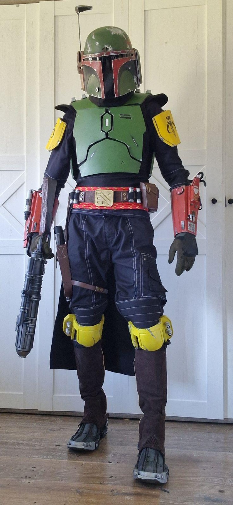 May include: A full-body costume featuring a green helmet with a T-shaped visor, green chest armor, yellow shoulder and knee plates, and red gauntlets. The outfit includes a black long-sleeved shirt, dark trousers, brown boots, and a blaster prop.