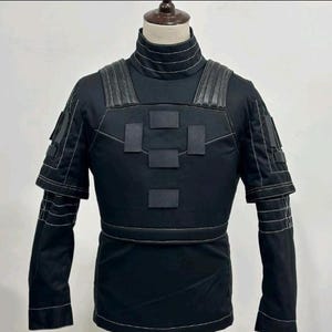 May include: A black tactical-style long-sleeve shirt featuring a high collar with horizontal stitching, structured shoulder panels, and a chest plate design with four rectangular patches. The garment includes white contrast stitching throughout.