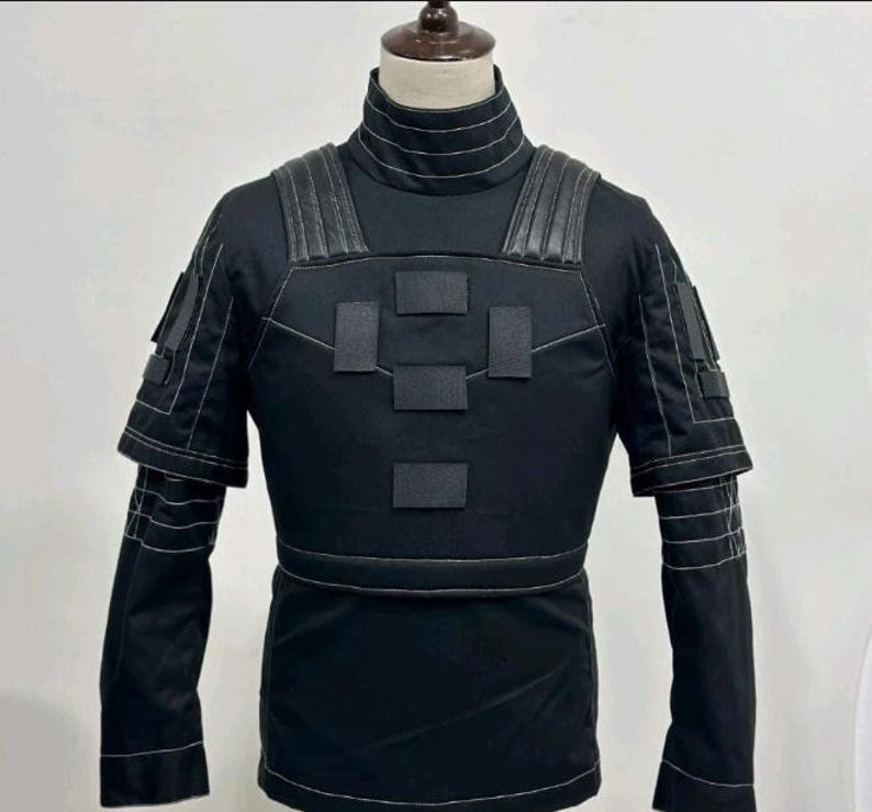 May include: A black tactical-style long-sleeve shirt featuring a high collar with horizontal stitching, structured shoulder panels, and a chest plate design with four rectangular patches. The garment includes white contrast stitching throughout.