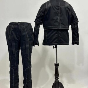 May include: A black two-piece outfit consisting of a jacket and pants. The jacket features a high collar, short sleeves, and white contrast stitching. The matching pants have a straight-leg cut, multiple panel details, and white contrast stitching.