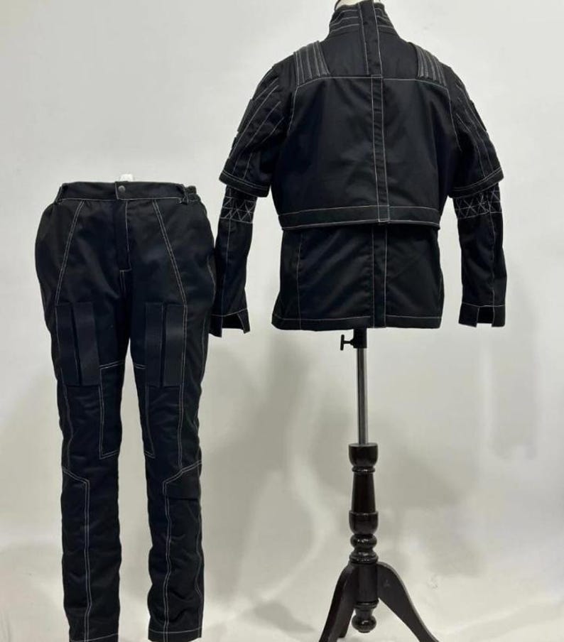 May include: A black two-piece outfit consisting of a jacket and pants. The jacket features a high collar, short sleeves, and white contrast stitching. The matching pants have a straight-leg cut, multiple panel details, and white contrast stitching.