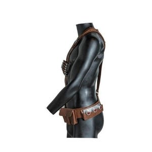 May include: A brown leather harness and belt set worn on a black mannequin torso. The harness features a chest-mounted ammunition holder, while the belt includes a rectangular pouch and three circular silver-toned metal accents with red centers.