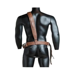 May include: A brown leather-like utility belt and shoulder strap harness worn across the torso. The set features a horizontal waist belt with attached metallic hardware components and a single vertical strap extending over the right shoulder.